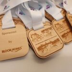 Wooden medals with white ribbons are laid out. Each medal is inscribed with "THE GREENHAM COMMON HALF AND 10k RUN" and sponsor details "BLOOR HOMES".