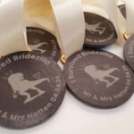 Close-up of multiple round medals with the text "I Survived Bridezilla," a silhouette of a dinosaur holding a bouquet, and the names "Mr & Mrs Hatten 04.04.24." White ribbons are attached to the medals.