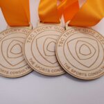 Three wooden medals with orange ribbons, engraved with "TSC Tricky Quiz 2023" and "The Sports Consultancy.