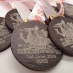 Close-up of several wooden medals for the Welsh Firefighter Challenge on June 1, 2024, featuring a dragon design and event name, with red-lettered ribbons attached.