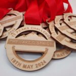 Wooden medals with red ribbons say "13 Peaks Challenge, The Lake District, 19th May 2024.