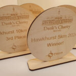 Wooden plaques for Dunk's Charity, showing awards for "Hawkhurst 10km 3rd Place" and "Hawkhurst 5km 2024 Winner!" with engraved building illustration.