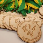 A pile of wooden medals, each engraved with "LOCKSBOTTOM CC PLAYER OF THE WEEK" and a horse image, with green and yellow ribbons attached.