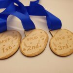 Three wooden medals with blue ribbons engraved with "SPUD 24" and labeled "1st," "2nd," and "3rd.