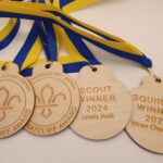 Four wooden medals with blue and yellow ribbons. Three are engraved with "Bill Ratcliff Award" and a fleur-de-lis. Two are labeled "Scout Winner 2024" and "Squirrel Winner 2024.