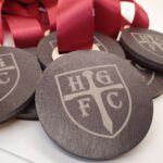 Close-up of black medals with "HG FC" logo and red ribbons on a white surface.