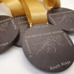 Three dark circular medals with gold ribbons, engraved with a mountain outline and labeled "Snowdon Four Ridges," "Crib Goch," "Lliwedd," "South Ridge," and "Y Liwedd.