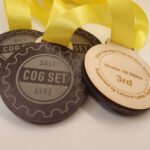 Three wooden medals with yellow ribbons, featuring "SALT COG SET AYRE" design. One medal reads "2024 Salt Ayre Cog Set Race Series, Under 10 open, 3rd.