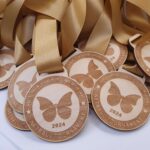 Wooden medals with a butterfly design and "Victoria Hawkins Memorial Netball Tournament 2024" text, attached to gold ribbons, displayed in a pile.