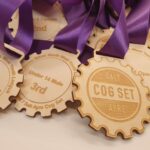 A close-up of wooden medals shaped like cogs with purple ribbons. Engravings on the medals show different categories, including "3rd Under 14 Male" and "Salt Ayre Cog Set.