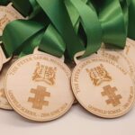 A pile of wooden medals with green ribbons, engraved with "The Peter Sakal Memorial Game, Gosfield School - 28th June 2024" and a coat of arms.