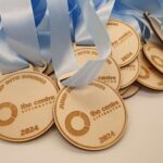 Wooden medals with light blue ribbons, inscribed with "JUMP INTO SUMMER, the centre Livingston, 2024," are shown in a pile.