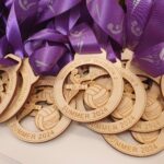 A pile of wooden medals with purple ribbons. The medals are engraved with "Winners of Division 2, Summer 2024, T.W.N.L, Netball.