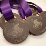 Close-up of medals with purple ribbons, featuring a cyclist engraved and text "Coast to Coast in a Day," dated 22/06/2024.