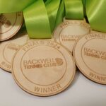 Wooden medals with green ribbons, engraved with "FINALS 2024 BACKWELL TENNIS CLUB WINNER," are shown in a close-up view.