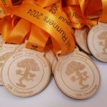 Wooden medals with "Alton Trail Runners 10K Challenge" and tree logo, attached to orange ribbons with "Trail Runners 2024" text.