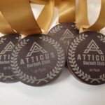 Three gold medals with ribbons are displayed. Each medal features the text Atticus Barbell Club 1st Place and a laurel wreath design surrounding the clubs logo.