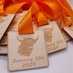 Wooden medals with orange ribbons are engraved with a highland cow image and the text Ormsary 10K 2024. Several medals are stacked in the background.