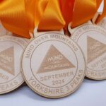 Wooden medals with orange ribbons are engraved with a triangle logo. Text reads Mind Over Mountains, Yorkshire 3 Peaks, and September 2024 on them. They are arranged in an overlapping pattern.