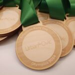 Wooden medals with green ribbons are stacked. Each medal is engraved with Employee Away Day, Urban & Civic, and Olympics 2024 in cursive script.