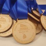 Close-up of several wooden medals with blue ribbons. The medals are engraved with Single-Handed Team Racing, NSSA, and Burton SC. They display various awards, such as Winner Silver Fleet 2024 and 2nd Place Gold Fleet 2024.