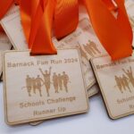 Wooden medals with orange ribbons are engraved with Barnack Fun Run 2024, Schools Challenge Runner Up, and a silhouette of runners. They are stacked together on a white surface.