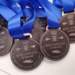 A collection of medals with blue ribbons, engraved with Royal Canoe Club Outrigger Hampton Court Challenge, featuring crossed paddles and a crown logo. The medals are stacked and partially overlapping on a smooth surface.