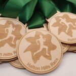 Wooden medals with green ribbons are stacked together. Each medal features a silhouette of a runner within a maple leaf design and the text Halton Hills Race.