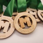 A close-up image of several wooden medals with green ribbons, engraved with MPC 24 Runner-Up and Midlands Pool Championship 24. The medals display a large M and text indicating SEMI-PRO.