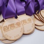 A pile of wooden medals with purple ribbons. The medals are engraved with EVT, Evesham Vale Triathletes, and SWIM - BIKE - RUN.