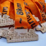 A pile of medals with wooden cutouts featuring the words 1st Shridge Canicrossers and an outline of a runner with a dog. The medals have orange ribbons printed with various distances like 8km and 20km.