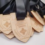 A pile of wooden medals with black ribbons. Each medal is shield-shaped, engraved with Inspire, Support, Educate, Northampton Saints Foundation, and the year 1880.