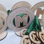 Wooden plaques and medals from the Midlands Pole Championships 2024 are displayed. The plaques are inscribed with WINNER, and the medals with RUNNER UP, each featuring a prominent letter M with green ribbons.