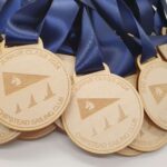 A pile of wooden medals with blue ribbons, engraved with CSC Junior Class 2024 and Chipstead Sailing Club. The medals feature a design of a sailboat and flag.