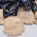 Wooden medals with a skull and compass design are stacked on a table. Each medal has a black lanyard and the text “Live Great Treasure Hunt” engraved on the front.