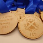 Wooden medals with blue ribbons are displayed. The engraved text reads 2024 Club Champs, 15 & Over, 1500m Free, Female, 2024 and 1st with an emblem featuring S.R.A. and a date founded 1896.