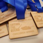 Wooden medals with blue ribbons engraved with Lifting the Decks 2024 and Challenge Winner, featuring an image of a tank and a crane.