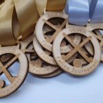 Wooden medals for the Fettes Tournament are displayed, featuring gold and silver ribbons. Each medal has crossed field hockey sticks with the words GOLD or SILVER engraved on top.