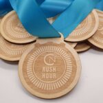 A close-up of several wooden medals with blue ribbons. Each medal features an engraved stopwatch design and the text Rush Hour. The medals are layered and spread out on a flat surface.