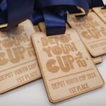 A close-up of several wooden medals with blue ribbons. Each medal is engraved with DEPOT YOUTH CUP 2024 and 1st PLACE. The medals are stacked and slightly overlapping on a light surface.