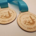 Two wooden medals with blue ribbons, engraved with 615 Trig Points & 1000 Miles 2004-2024 and a lighthouse design.