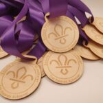 A pile of wooden medals adorned with purple ribbons. The medals feature a fleur-de-lis symbol and are engraved with Scouts Hereford and Worcester Top Awards Achiever.