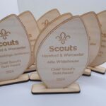 Wooden Scout awards with engraved fleur-de-lis symbol, labeled Scouts Hereford & Worcester Chief Scouts Gold Award 2024, featuring names like Oliver Seekings-Jenkinson and Alfie Whitehouse, arranged in a fan layout.