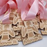 A stack of wooden medals with pink ribbons. Each medal features a silhouette of a person in a dynamic pose and the words Move It engraved below.