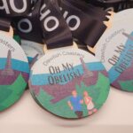 Close-up of medals with Dawlish Coasters Oh My Obelisk! text. Features an illustration of a landscape with two runners and an obelisk in the background. Black ribbon attached to each medal.