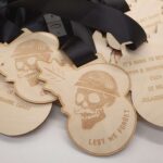 A stack of wooden medals with black ribbons. The medals are shaped like skulls wearing helmets, engraved with Lest We Forget. Text on one visible medal includes 2024, 22 miles, and Jurassic Coast.