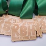 Wooden medals with green ribbons, engraved with KBH LINE DRIVE QUALIFYING RUN and an image of a runner.