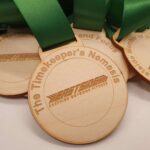 Close-up of wooden medals with green ribbons displaying engraved text: Most Determined and Focus, The Timekeepers Nemesis, Eastside Outdoor Fitness, and Member of the Year in the background.