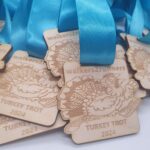 A close-up of several wooden medals with blue ribbons. The medals feature an engraved image of a turkey and are labeled walkers2runners TURKEY TROT 2024.