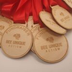 A pile of wooden medals with red ribbons. The medals are engraved with FUNDRAISER and BEE UNIQUE AUTISM, along with a bee design.